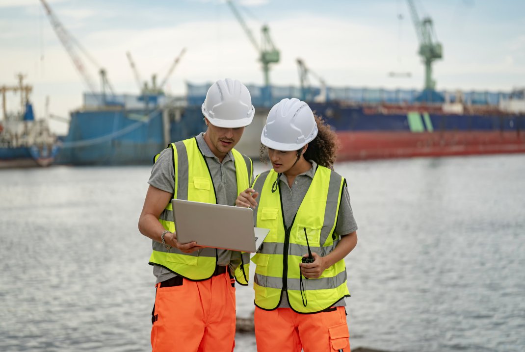Two technicians in hi-vis safety vests and hard hats reviewing a project on a laptop on site